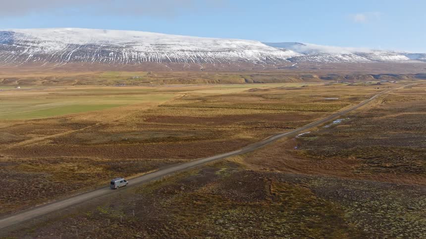 An aerial shot follows a camper van driving down a long, straight dirt road through a vast, barren valley toward a range of snow-capped mountains in Iceland