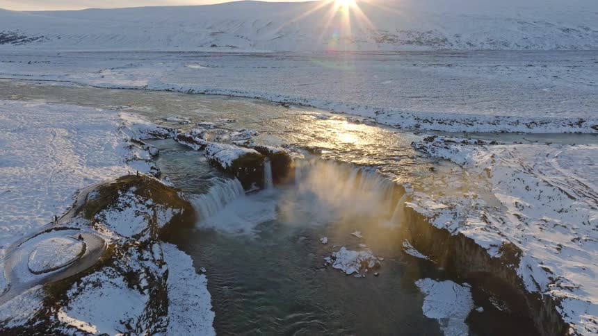 A breathtaking aerial shot pulls back to reveal the powerful Godafoss waterfall in a vast, snow-covered Icelandic landscape at sunset