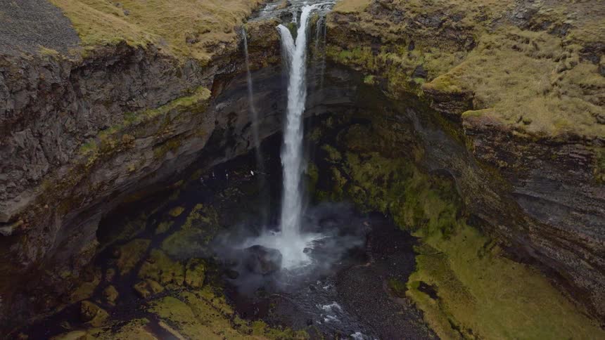 A stunning aerial shot pulls back to reveal the powerful Kvernufoss waterfall plunging into a mossy, rocky canyon in Iceland