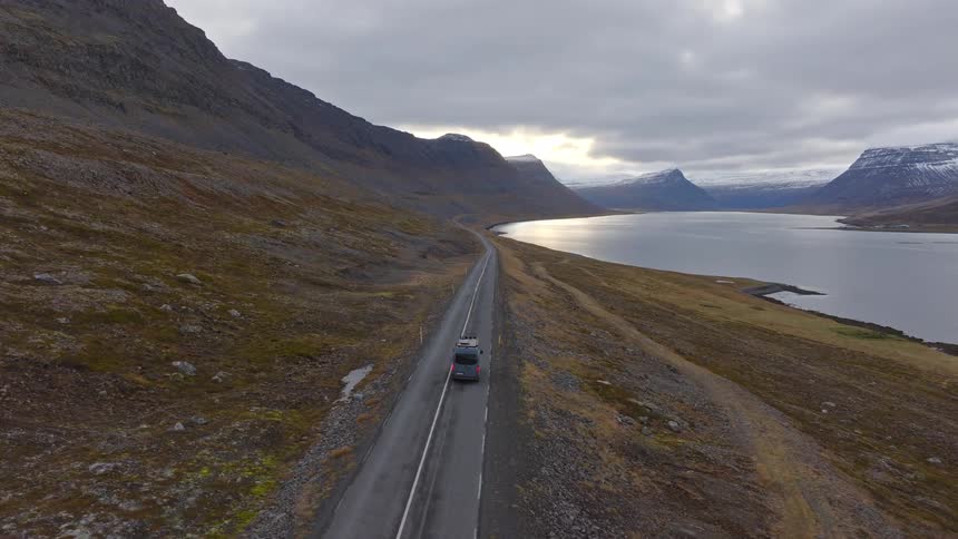 Aerial view of a camper van driving on a remote road alongside a fjord, surrounded by a dramatic mountainous landscape in Iceland