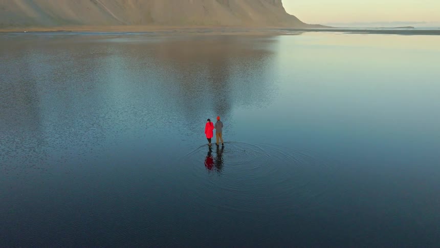 An epic aerial pullback reveals a couple standing together in the perfectly still, reflective waters of a tidal lagoon at the base of the majestic Vestrahorn mountain in Iceland
