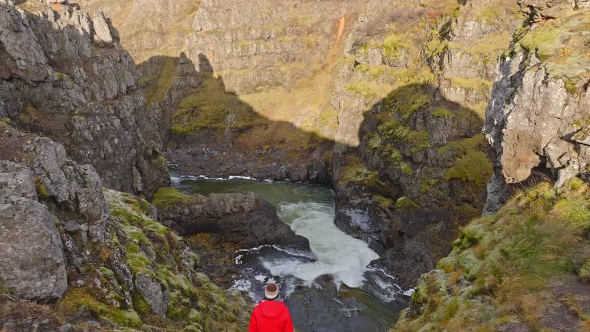 A dramatic aerial view pulls back from a rushing river inside a deep, mossy canyon to reveal a lone traveler in a red coat on the cliff's edge