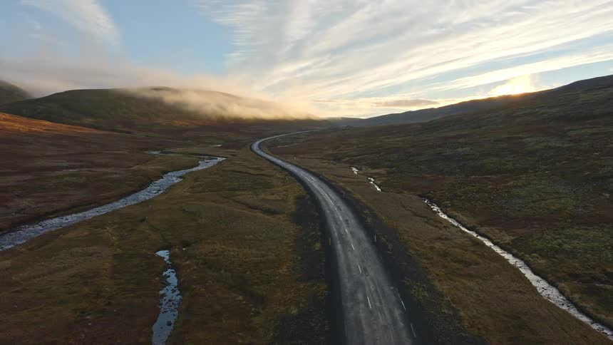 An aerial shot flies backwards over an empty winding road through a vast valley with foggy mountains at sunrise