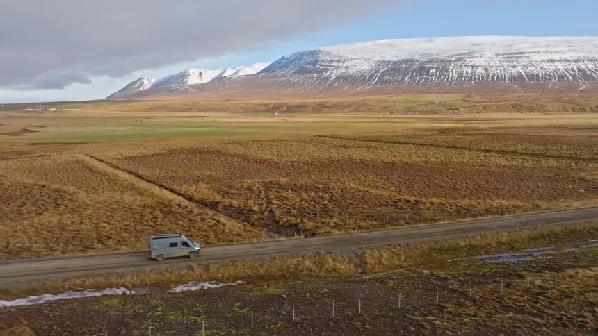 Aerial view of a camper van driving along a remote dirt road through the expansive Icelandic countryside with snow-capped mountains in the background