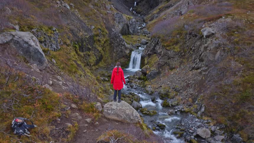 Aerial view of a woman in a red jacket hiking and standing on a rock to admire a majestic waterfall in a remote mountain canyon