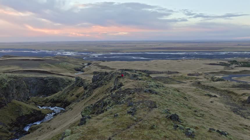 Aerial shot rises over a rocky Icelandic canyon at sunset, revealing a person in a red coat admiring the vast river valley below