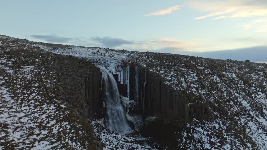 Majestic aerial view pulls back to reveal a powerful waterfall cascading over unique basalt rock columns in a snow-covered winter landscape