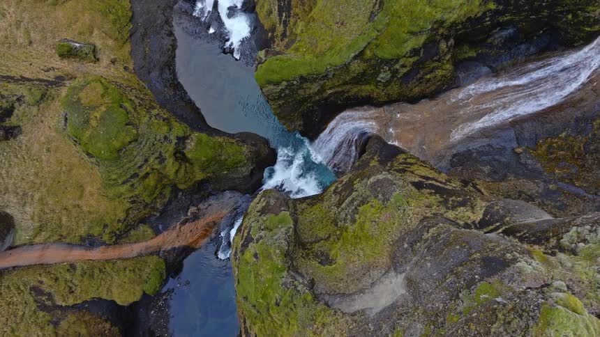 A stunning top-down aerial shot starts on a turquoise waterfall before ascending to reveal a vast, deep canyon with moss-covered cliffs