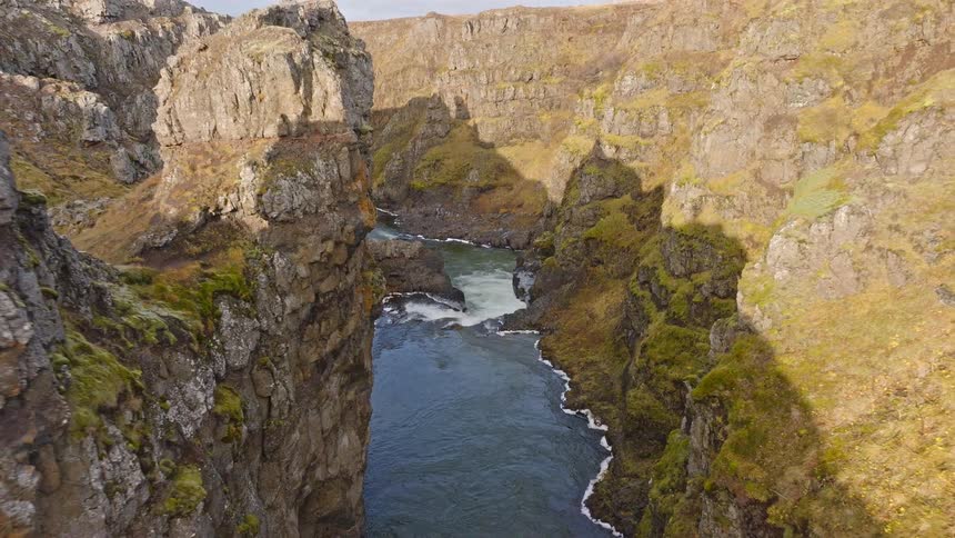 A slow aerial pull-back reveals a rushing river carving its way through a deep, rocky, moss-covered canyon in the Icelandic highlands