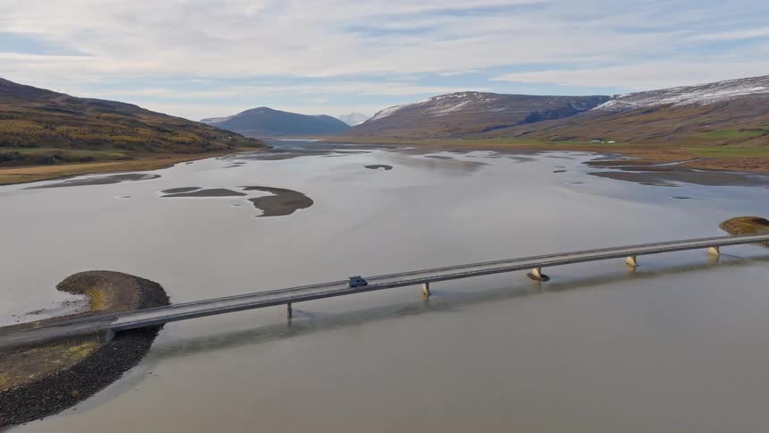 A lone car takes a scenic drive across a long bridge over a wide river valley with snow-capped mountains in the distance