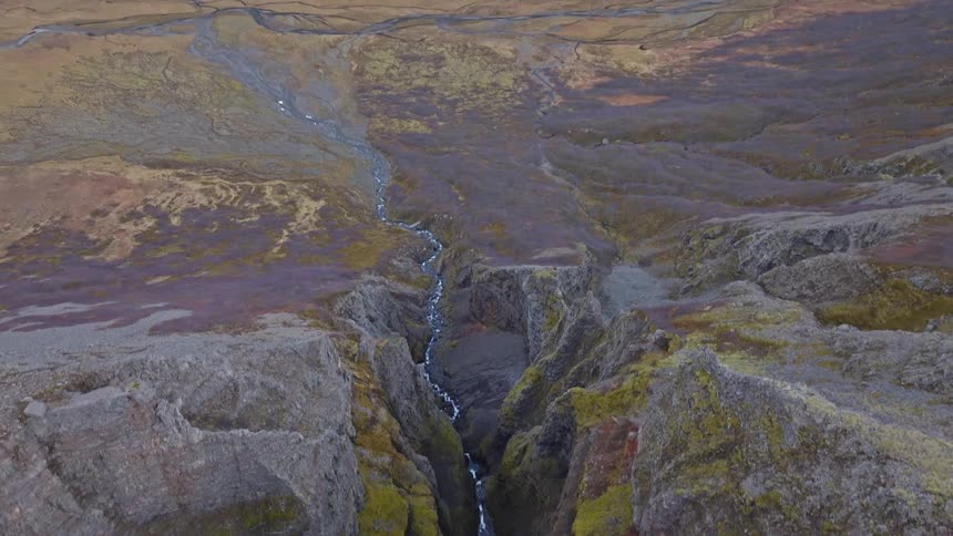 A dramatic aerial reveal shot pulls back from a deep river canyon to showcase a vast, rugged valley and snow-dusted mountain range