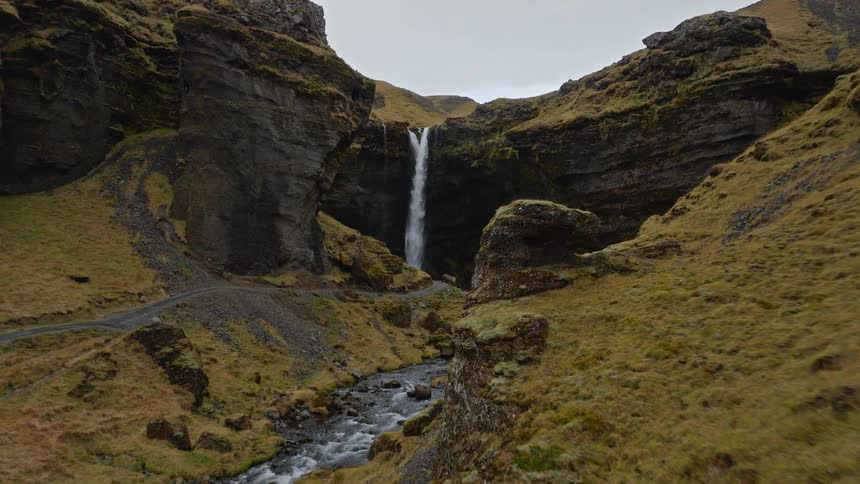 An aerial drone shot flies past a hiker in a red jacket, revealing a stunning waterfall in a remote Icelandic canyon