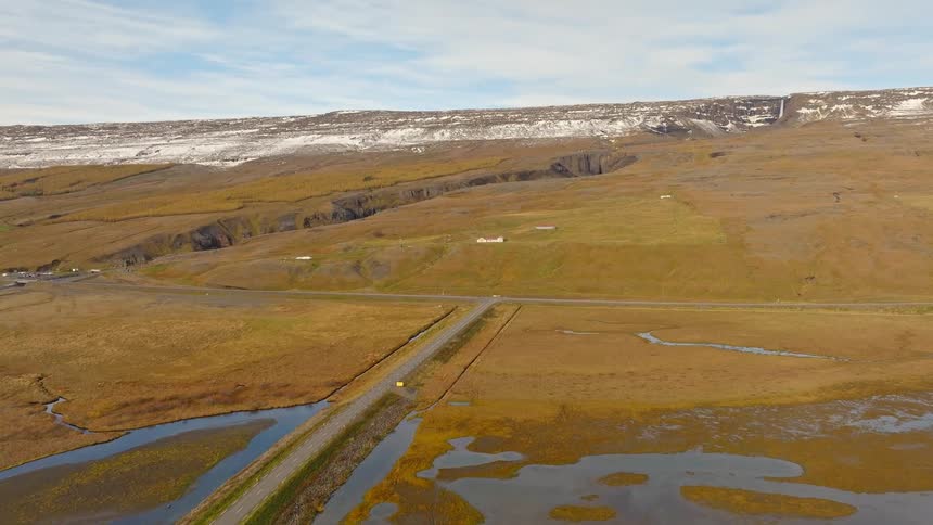 A breathtaking aerial view follows a car driving along a highway through a vast Icelandic wetland toward distant snow-capped mountains