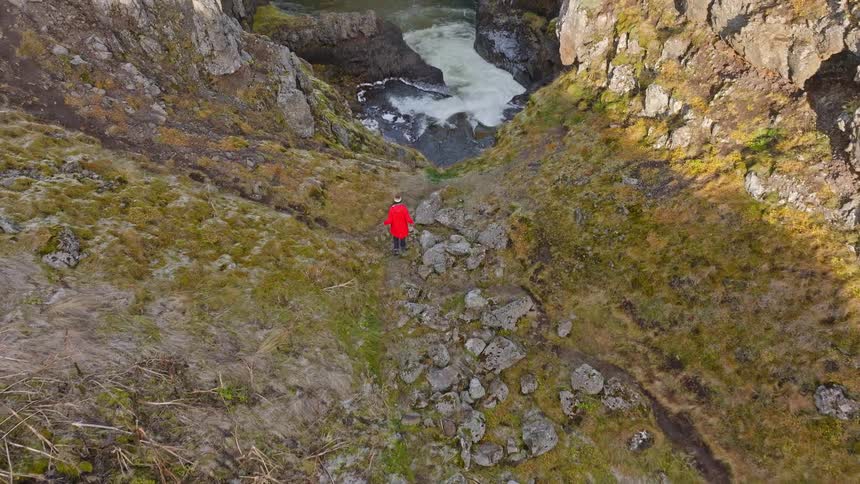 An aerial view follows a person in a bright red coat hiking down a rugged, winding trail to the edge of a sunlit canyon with a rushing river below