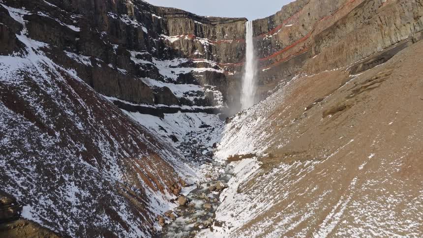 An aerial view flies up a snowy, rocky river canyon to reveal the magnificent Hengifoss waterfall in Iceland