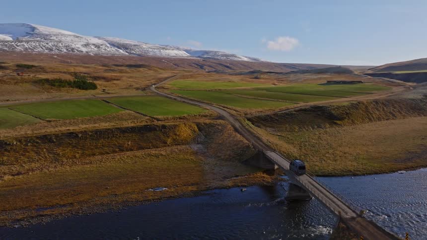 An aerial shot follows a camper van driving across a bridge over a river, revealing a vast Icelandic landscape with snow-capped mountains