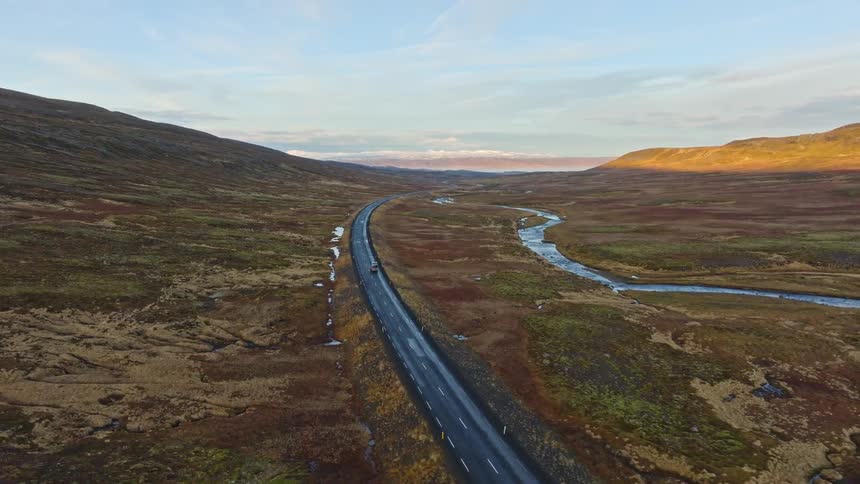 An aerial view captures cars driving on a scenic, winding road through a vast, remote valley alongside a river during golden hour