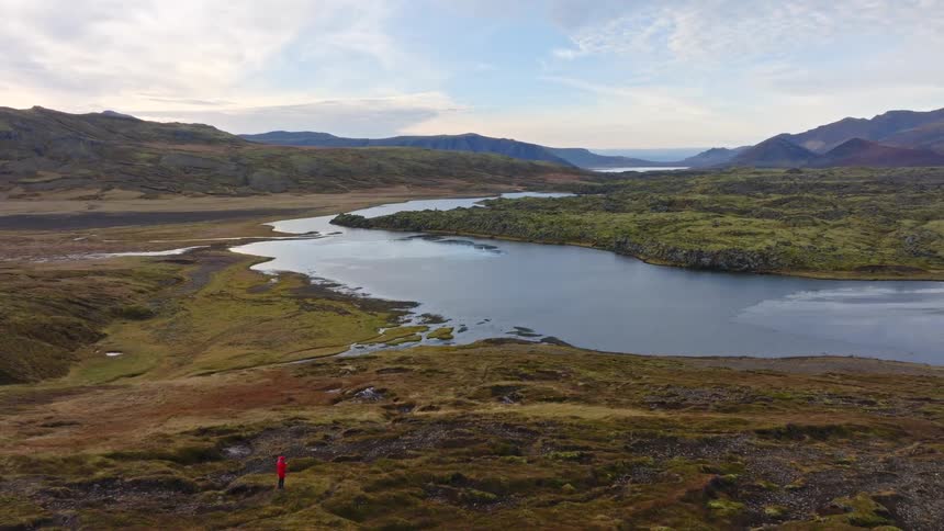 An aerial pan reveals a solitary figure in a red jacket standing amidst the vast, rugged Icelandic landscape, overlooking a tranquil lake and distant mountains