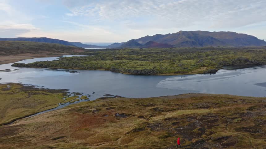 An aerial pan reveals a lone hiker in a red jacket walking through a vast and epic volcanic landscape next to a lake in Iceland