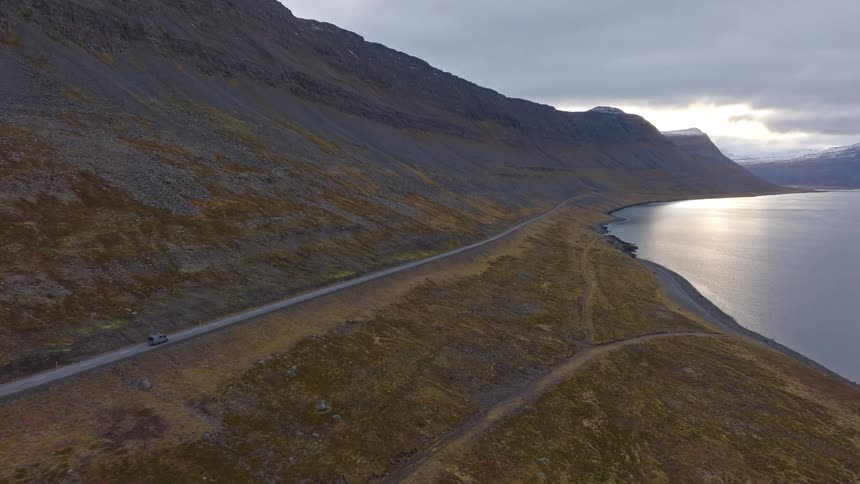 An aerial view of a car driving along a remote coastal road beside a fjord at the base of a rugged mountain range in Iceland