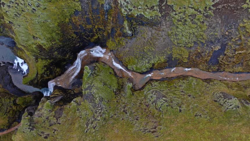 A slow aerial ascent reveals a spectacular river carving its way through a vibrant green, mossy canyon in Iceland