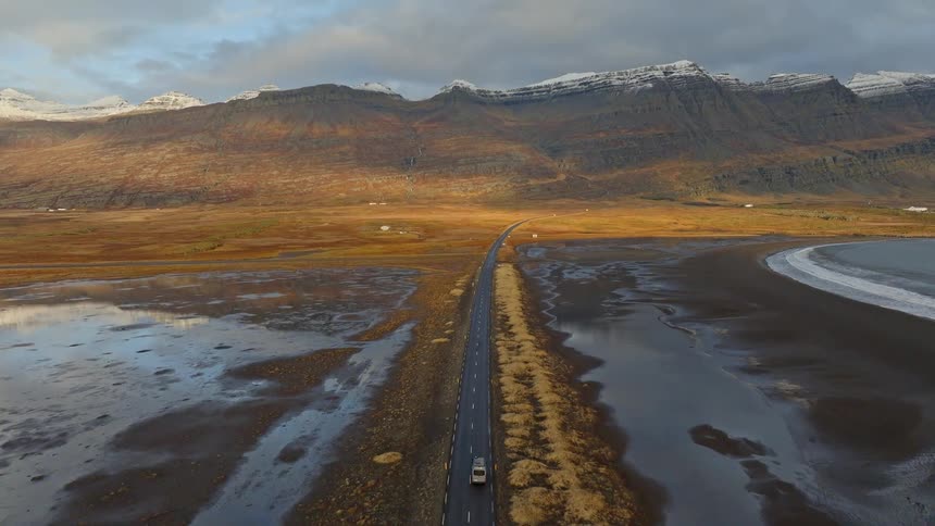 An aerial view shows a camper van on a road trip, driving along a remote highway towards snow-capped mountains in a vast Icelandic landscape