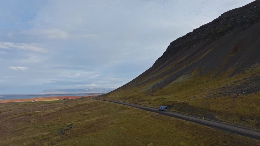 An aerial view captures a campervan on a scenic road trip through the vast Icelandic countryside, driving alongside a dramatic mountain