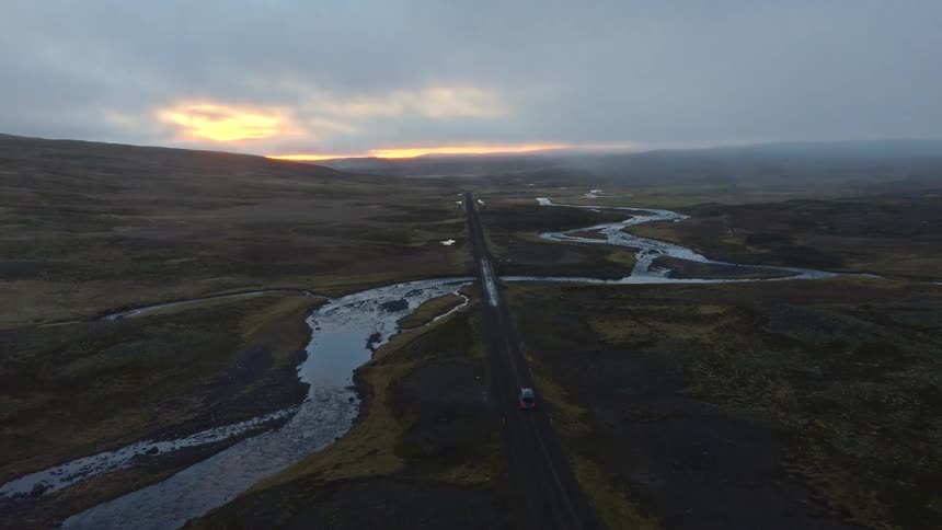 An aerial view follows a lone car driving down a remote highway through a vast, rugged landscape next to a meandering river during a dramatic sunset