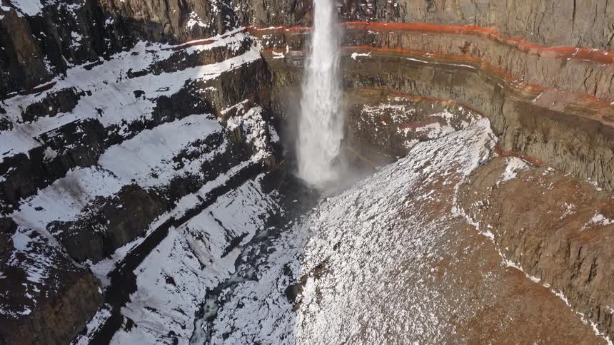 An aerial view tilts up from a snowy river canyon to reveal the powerful Hengifoss waterfall cascading over dramatic, colorful rock layers in Iceland