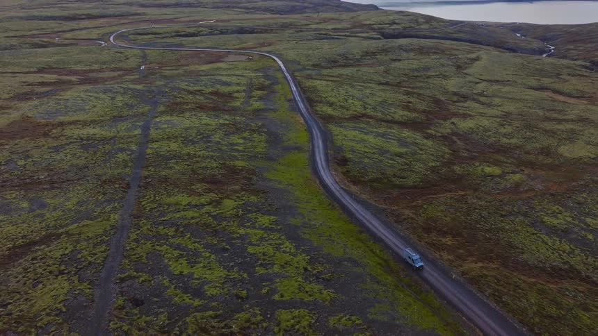 An aerial view follows a single vehicle driving along a remote dirt road through a vast, moss-covered landscape in Iceland