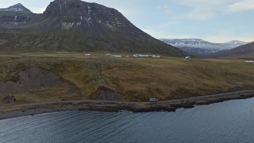 A camper van travels along a winding coastal road next to a calm fjord, surrounded by majestic snow-capped mountains