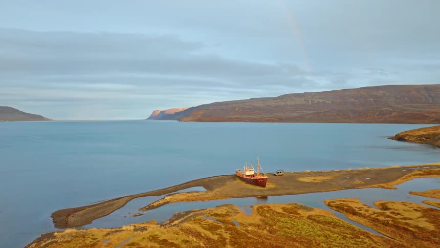 An aerial view pulls away from a beached shipwreck in Iceland, revealing the vast, beautiful fjord and a rainbow in the distance