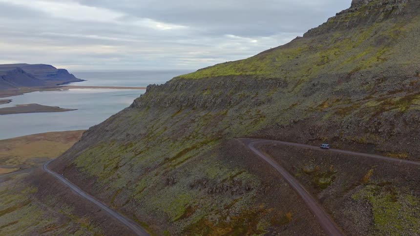 An aerial view shows a camper van navigating a winding dirt road up a rugged, mossy mountain in the vast, remote Icelandic Westfjords