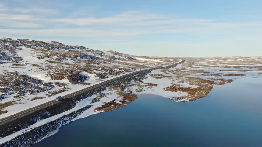 Aerial footage tracks a camper van driving along a scenic road next to a calm fjord in a vast, snow-covered Icelandic landscape