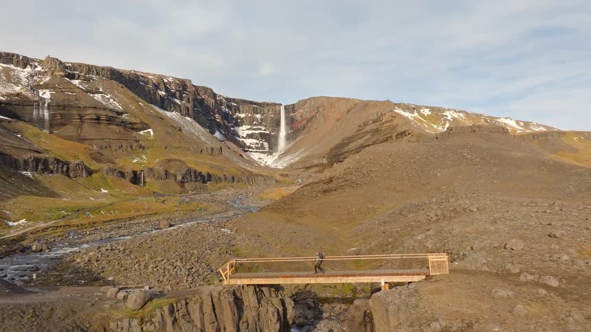 Aerial pullback shot shows a solo hiker crossing a modern bridge over a deep canyon, revealing the epic Hengifoss waterfall in the Icelandic highlands