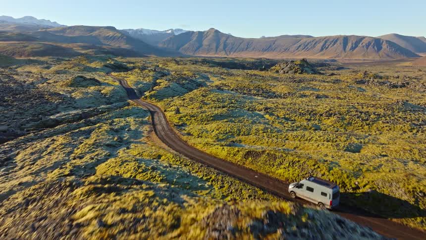 Aerial footage follows a camper van as it drives along a scenic dirt road through Iceland's vast, mossy lava fields towards distant mountains