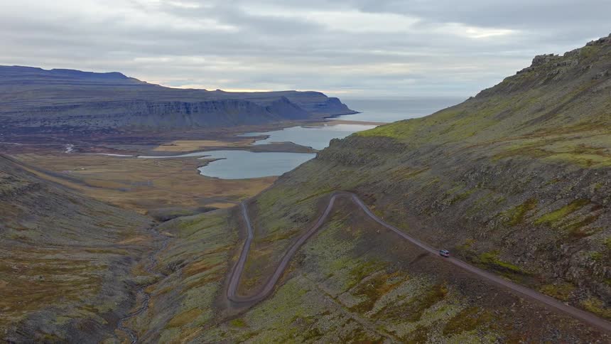 Aerial view of a car driving on a winding dirt road through a rugged Icelandic mountain pass overlooking a fjord and the distant ocean