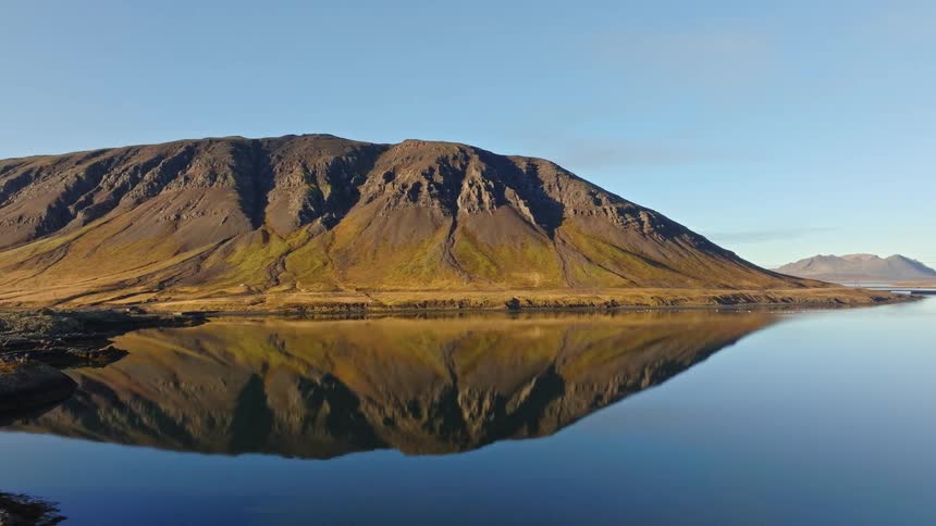 Aerial view of a majestic Icelandic mountain perfectly reflecting in the calm, glassy waters of a fjord on a sunny day