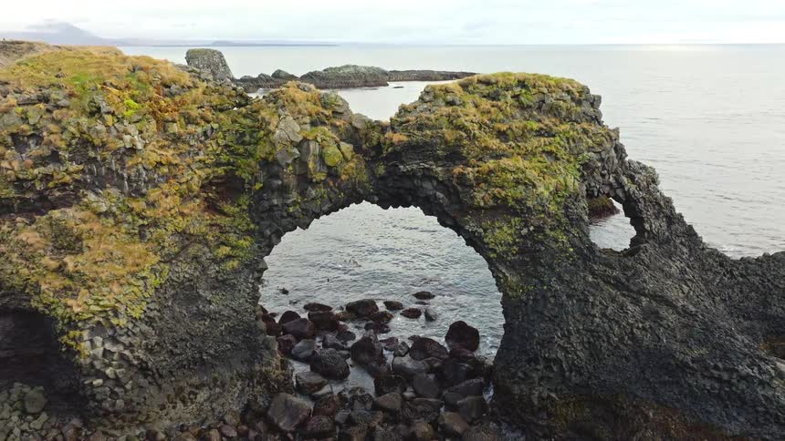 An aerial drone shot slowly pulls back to reveal a magnificent moss-covered volcanic rock arch on the rugged coastline of Iceland