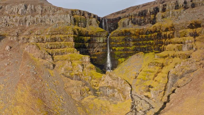 Drone pulls back to reveal a majestic waterfall flowing down a series of tiered, mossy cliffs in a vast, rugged canyon