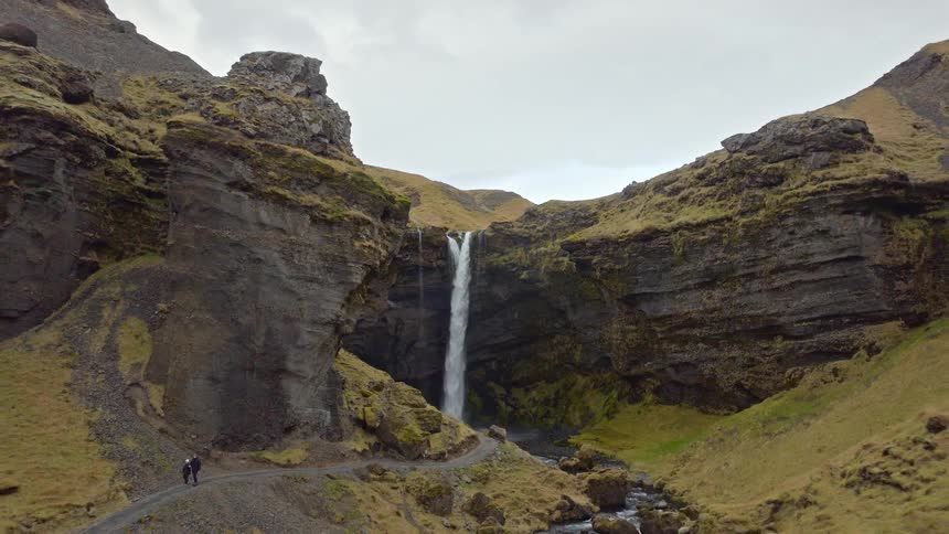 A drone pulls back from a stunning waterfall in a rocky Icelandic canyon to reveal its immense scale and the people admiring its beauty