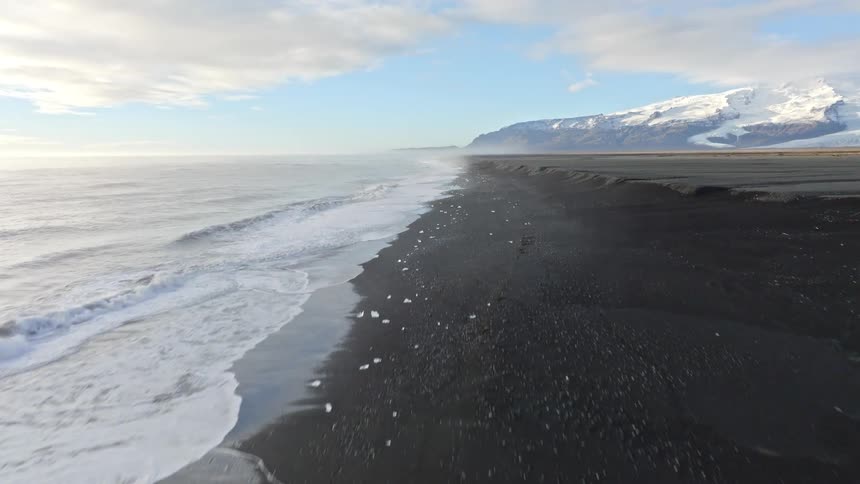 Aerial view flying along the dramatic shoreline of Iceland's black sand Diamond Beach with ice chunks and distant snowy mountains