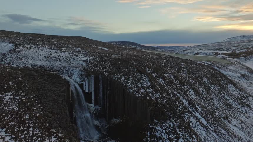 An aerial view pulls back from a stunning winter waterfall over basalt columns to reveal a vast, snowy valley at sunrise