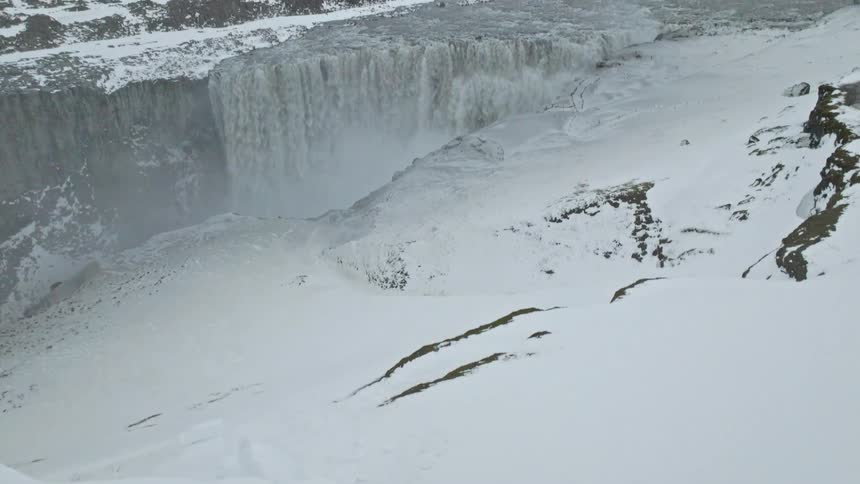 A stunning aerial shot pans from a lone person on a cliff to reveal the majestic and powerful Dettifoss waterfall in a vast, snowy Icelandic landscape
