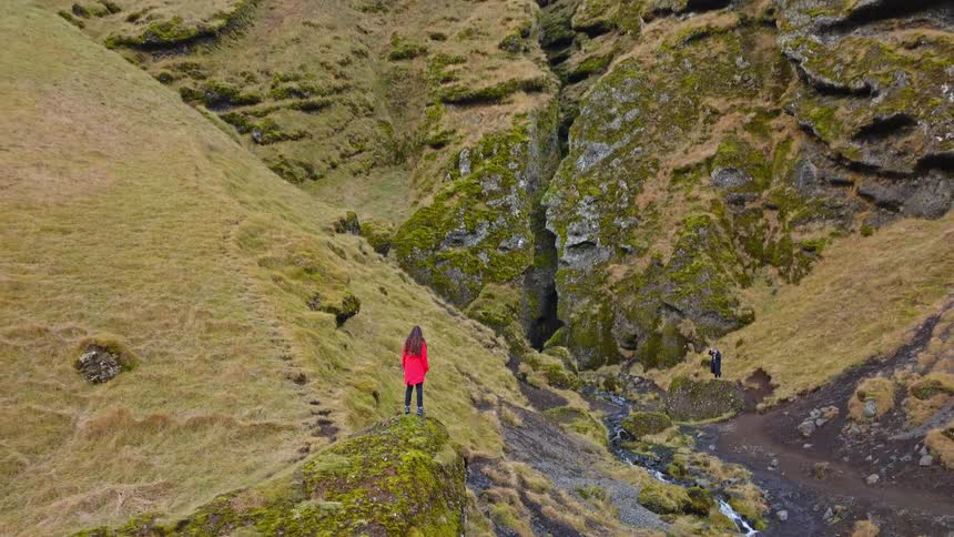 Aerial view slowly reveals a majestic green mossy canyon in Iceland as a woman in a red coat admires the epic view
