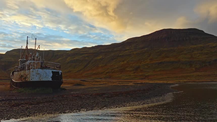 An old rusty shipwreck lies abandoned on the shore of a vast, mountainous fjord in Iceland during a golden sunset