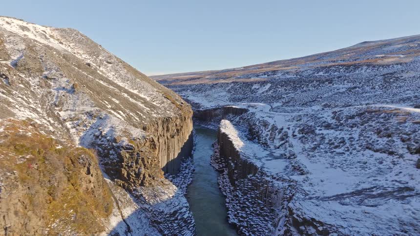 A breathtaking aerial flight follows a winding river through a majestic, snow-covered canyon with unique basalt column walls in Iceland