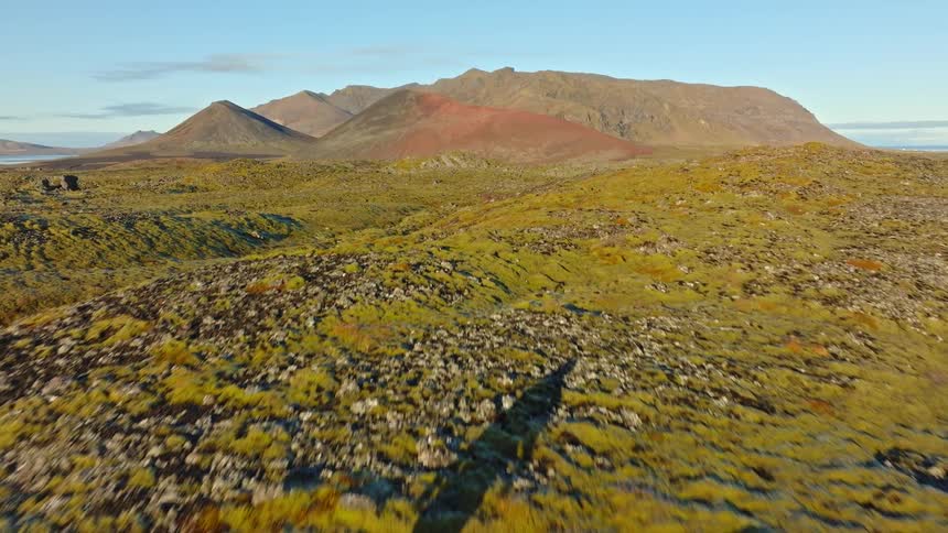 An aerial view follows a lone hiker in a red jacket exploring a vast, moss-covered volcanic landscape under a clear sky