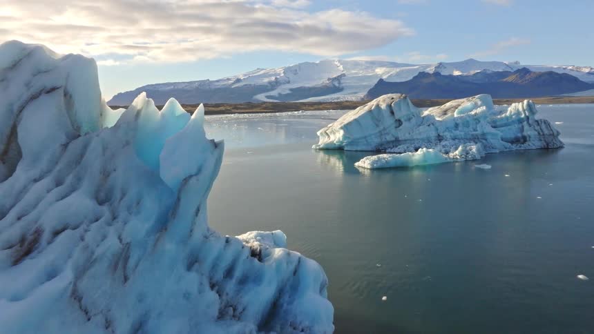 A drone flies slowly past majestic blue icebergs floating in a tranquil glacial lagoon in Iceland