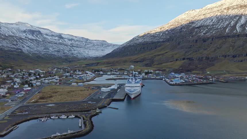 Stunning aerial pullback reveals a large ferry docked in a small harbor town nestled within a majestic, snow-capped mountain valley